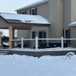 Covered backyard deck with white railing, natural wood posts, and snow-covered roof beside a two-story home.