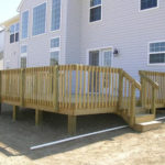 Pressure-treated wood deck with railing and stairs attached to the back of a two-story home.