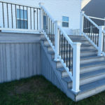 Elevated gray deck with wide stairs, white railing, and black balusters beside a backyard.