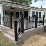 Covered backyard deck with gray composite boards, black railing, and steps leading to a patio area.