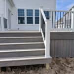 Gray backyard deck with wide stairs, white railing, and black balusters attached to a two-story home.