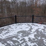 Gray composite deck with black metal railing and patches of snow overlooking a wooded backyard.
