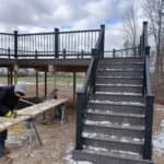Elevated gray deck with black railings and staircase during winter installation beside a house.