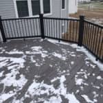 Gray composite deck with black metal railing and light snow beside a two-story house.