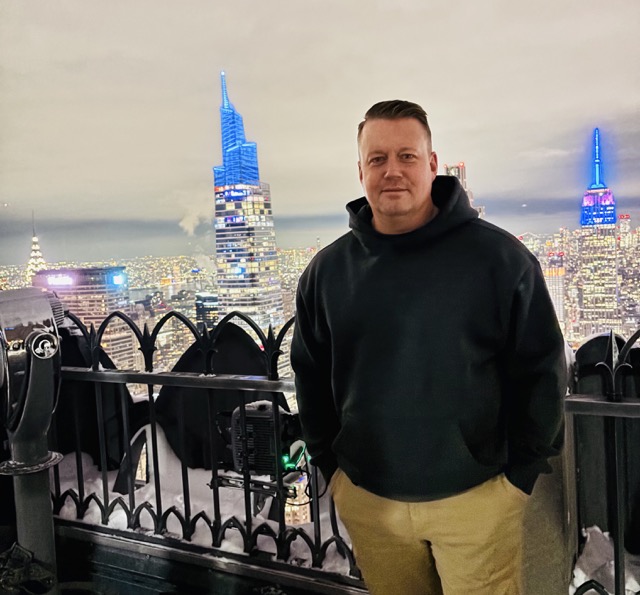 A person in a black hoodie and tan pants stands on an outdoor observation deck at night, overlooking a brightly lit city skyline with colorful illuminated skyscrapers in the background.