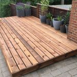 Alt Text A low-profile natural wood platform deck installed over a stone paver patio next to a brick building, decorated with several large potted plants.