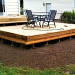 Alt Text A low-profile wood platform deck with built-in stairs, featuring a dining table and chairs set up in a backyard area with fresh dirt ground.