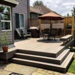 A raised backyard deck attached to a gray house, featuring wide steps, built-in bench seating, Adirondack chairs, a dining set with an umbrella, and a wooden privacy screen.