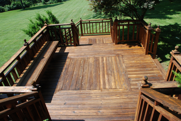 A multi-level wood deck featuring built-in perimeter bench seating, wood railings with decorative post caps, and a step-down platform, set against a grass lawn and trees.