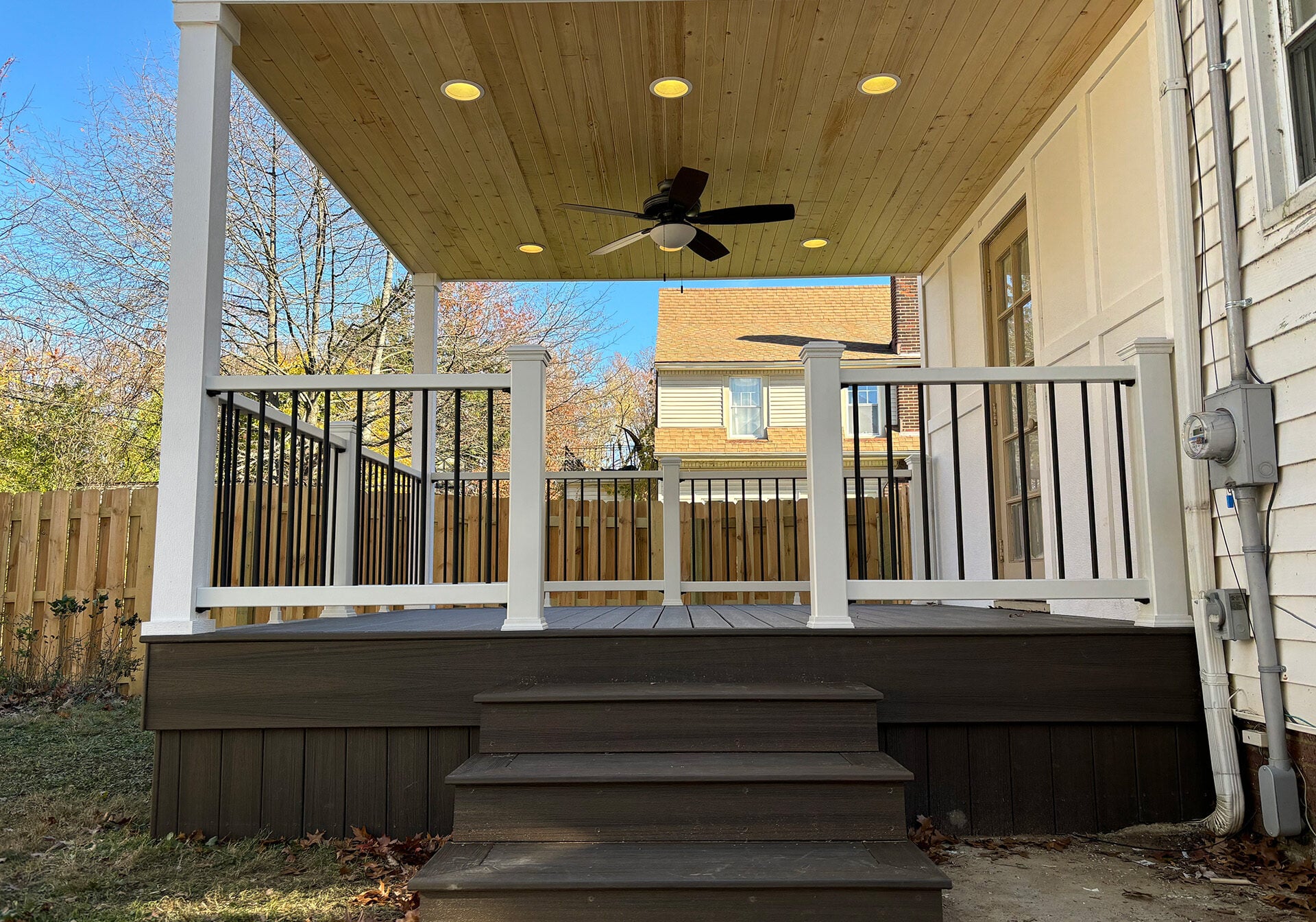 Covered backyard deck with dark composite boards, white railing, wood ceiling, recessed lights, and ceiling fan.