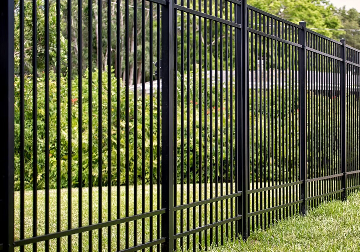 A black aluminum ornamental fence featuring vertical pickets and a lower horizontal rail, installed on a green lawn with lush bushes in the background.