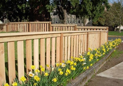 A natural wood fence featuring vertical slats and a flat top rail bordering a garden bed with yellow daffodils.